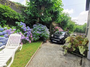 A garden with a car parked in front of a building, a white plastic chair, and various flowering plants.
