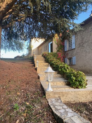Una escalera de piedra que conduce a un edificio con contraventanas azules, rodeado de vegetación y un cielo despejado.