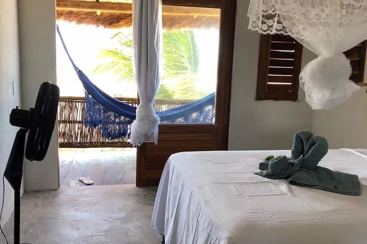 Ein Zimmer mit Bett, Ventilator, Hängematte und Blick auf Palmen.