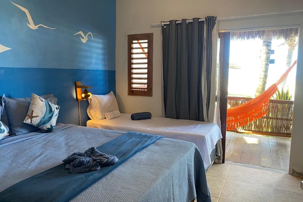Ein Hotelzimmer mit zwei Betten, einem Fenster mit Jalousien, einer Hängematte draußen und einer Wand mit Möwenmotiven.