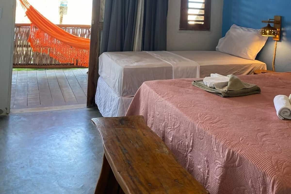 Ein Zimmer mit zwei Betten, einer Bank, einem Fenster mit Jalousien, einer Pendelleuchte und Blick auf eine Palme draußen.