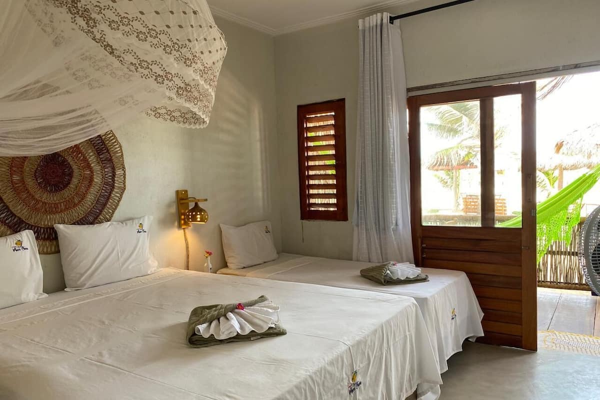 Ein Zimmer mit zwei Betten, einer großen gewebten Wandteppich, einer Wandleuchte und Blick auf eine tropische Außenanlage durch eine Schiebetür.