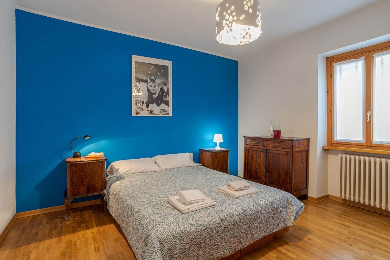 A bedroom with a blue wall, a bed with a patterned bedspread, two bedside tables with lamps, a wooden cabinet, and a window with a view.
