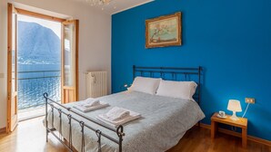 Un dormitorio con cama, mesita de noche, lámpara, un cuadro en la pared y vista al mar a través de una ventana.