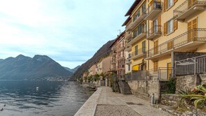 Un hotel frente al agua con balcones, un camino de piedra y montañas al fondo.