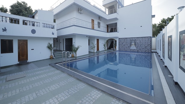 A modern building with a swimming pool, multiple balconies, and a tiled walkway. - APK Farm House (Noida)