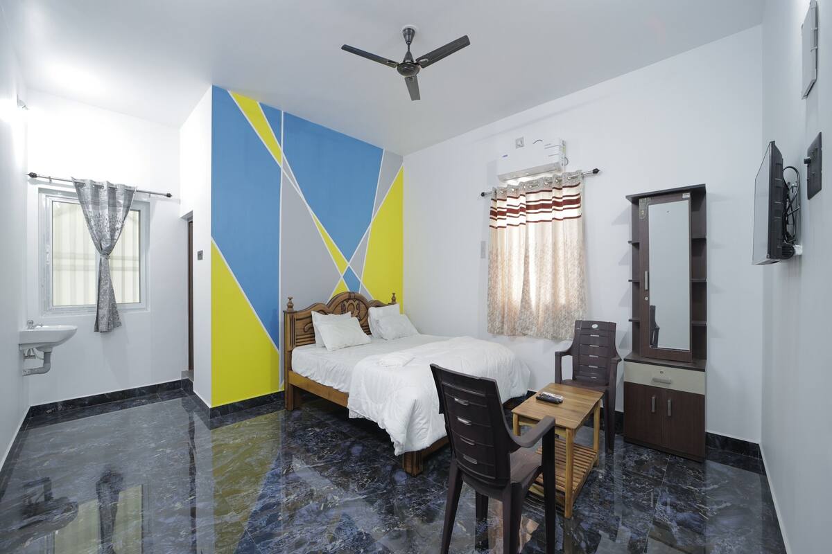 Quarto de hotel com cama, escrivaninha, cadeira, espelho, ventoinha no teto e banheiro com pia e janela.