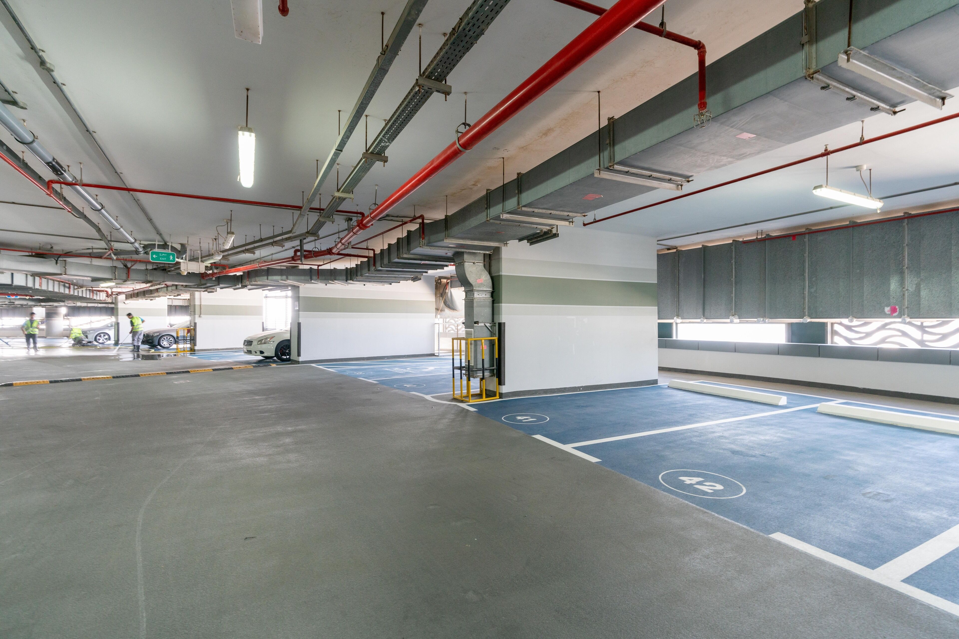 A parking garage with blue parking spaces, visible pipes, and a yellow safety barrier.