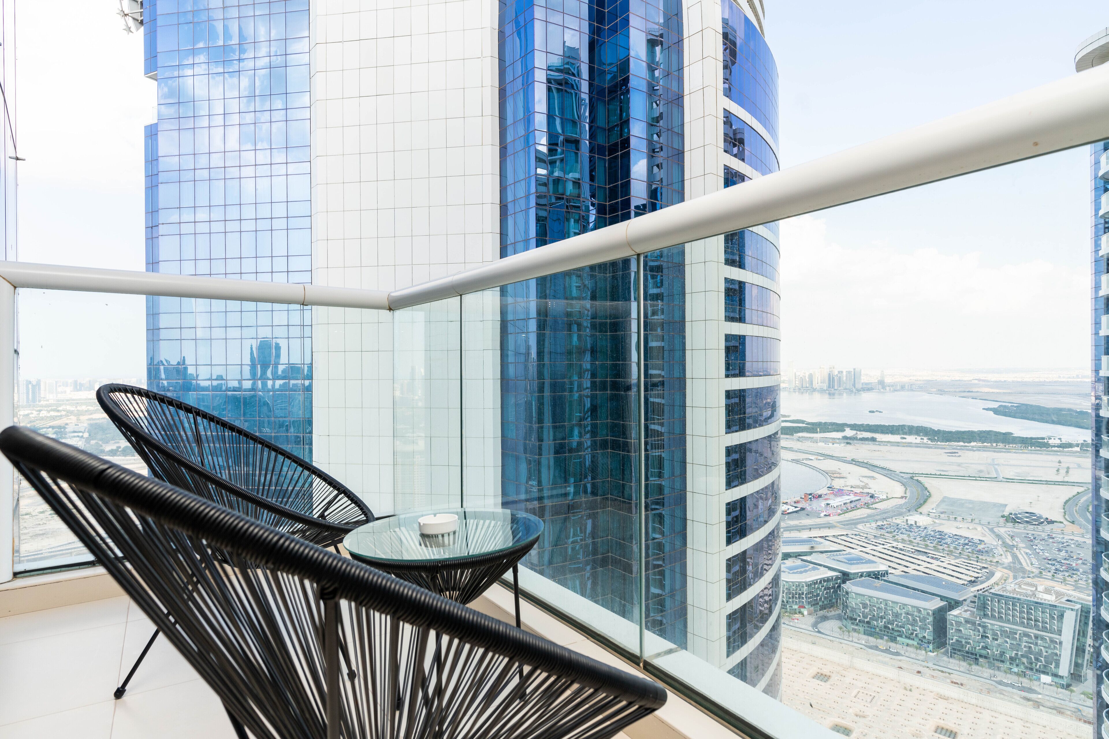 A balcony with a glass table and two chairs, overlooking a cityscape with modern buildings and a river.
