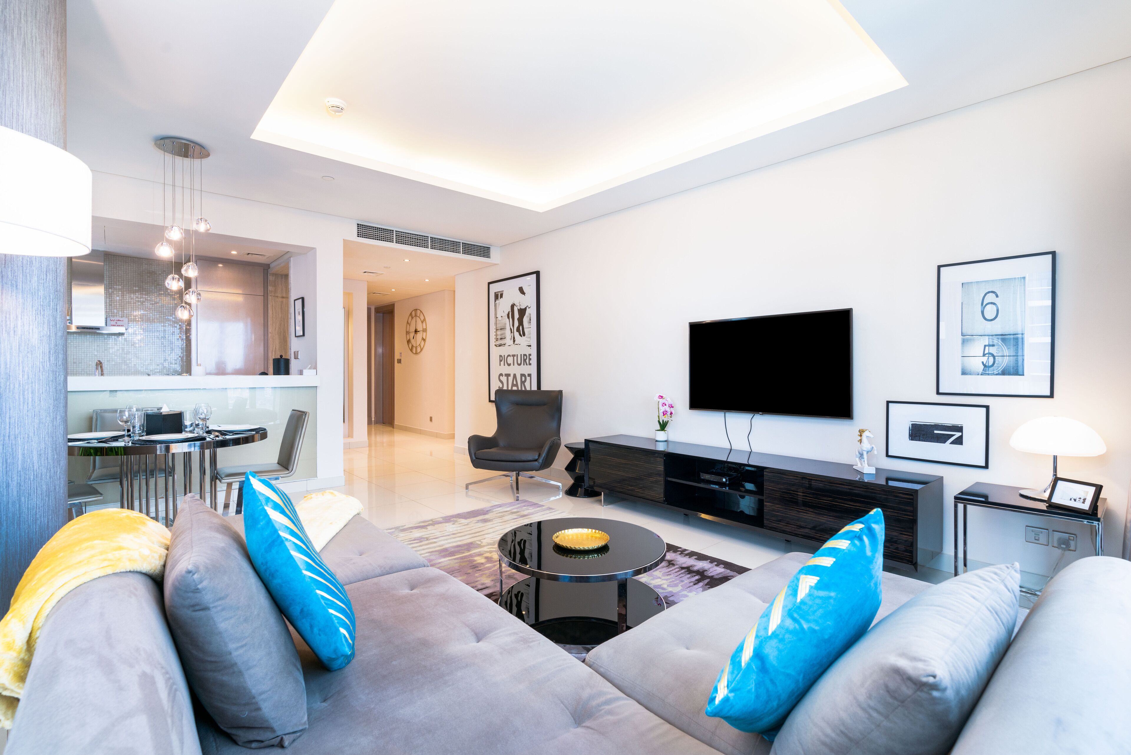 A modern living room with a sofa, coffee table, and a flat-screen TV. There is a dining area with a table and chairs, and a kitchen in the background.