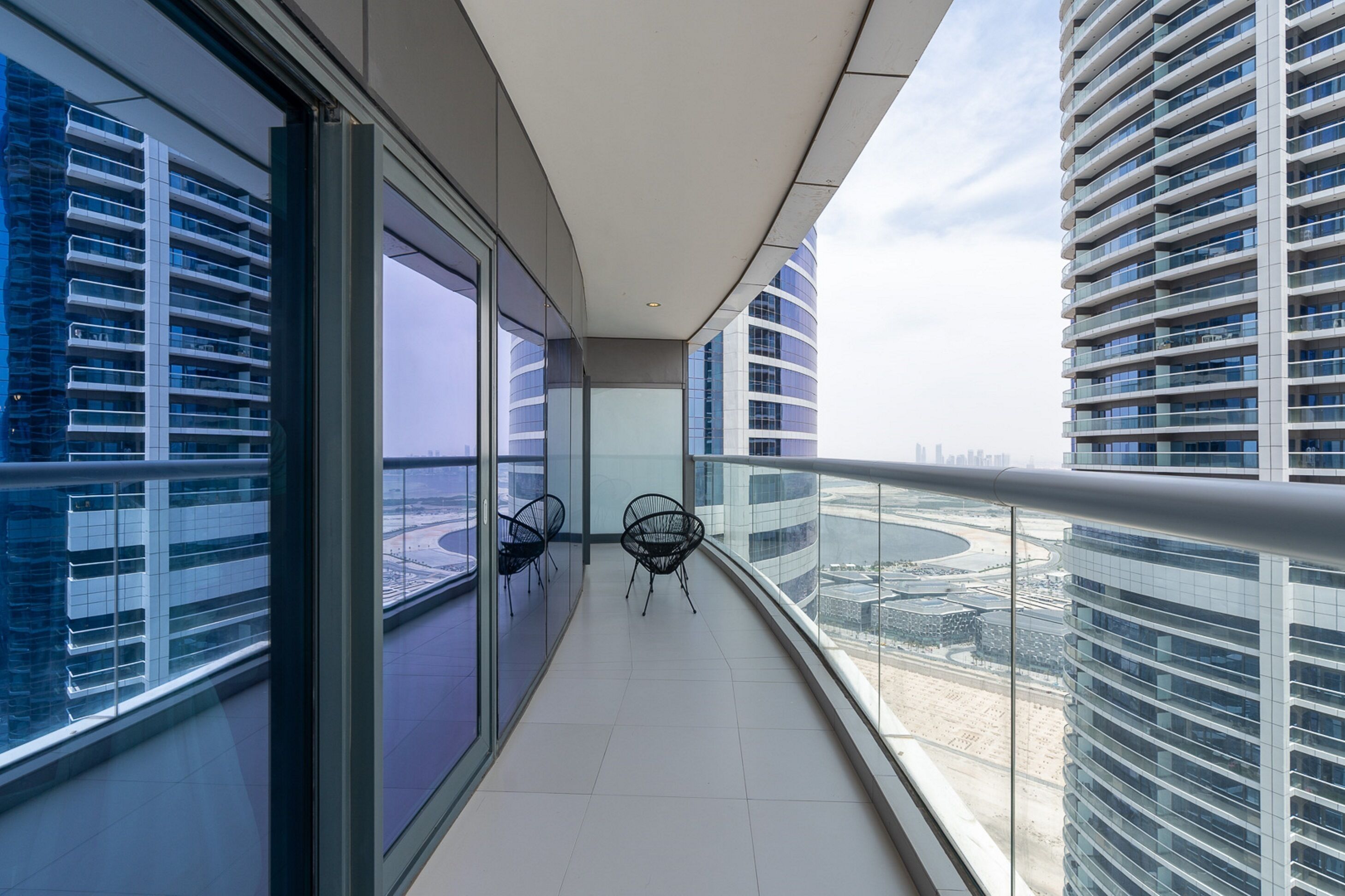 A modern balcony with glass railings, a view of a cityscape, and two chairs.