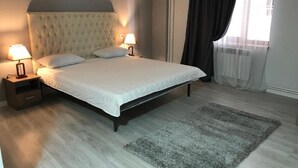 A bedroom with a tufted headboard, two bedside tables with lamps, a large bed, a grey rug, and a window with curtains. - The Friends Hotel (Yerevan)
