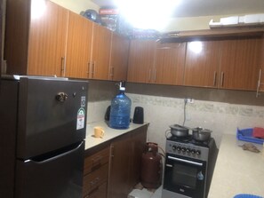 A kitchen with wooden cabinets, a refrigerator, a large water dispenser, a gas cylinder, a stove, and a coffee maker. - Family sized condo (Nairobi)