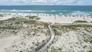Una playa con un paseo marítimo de madera que lleva a la orilla, rodeada de dunas y sombrillas de playa.