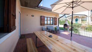 An outdoor patio with a wooden table and chairs, a large umbrella, and a view of a building with a green door. - Residenza Airone (Polpenazze del Garda)