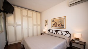 A bedroom with a bed, a wardrobe, a nightstand with a lamp, a painting on the wall, and an air conditioning unit. - Residenza Airone (Polpenazze del Garda)