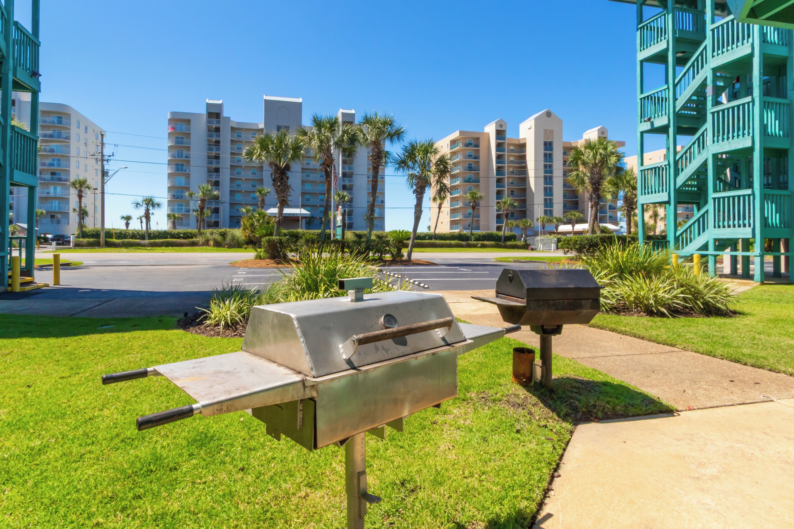 A grassy area with a barbecue grill and a trash can, surrounded by palm trees and a parking lot.
