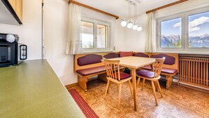 Un comedor con mesa y bancos de madera, una cocina con estufa y armarios, y ventanas amplias con vista a la montaña.
