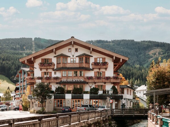 Un hôtel de style alpin traditionnel, avec des balcons en bois ornés de jardinières fleuries, une enseigne portant le nom « HUBERTUS » et une vue sur une chaîne de montagnes en arrière-plan.