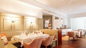 A restaurant with a wooden floor, white tables, and chairs. There is a counter with a vase of flowers and a painting. - Geniesserhotel Hubertus (Filzmoos)