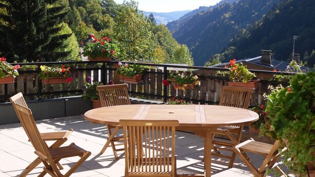 Une terrasse extérieure avec une table ronde en bois et des chaises, entourée de plantes en pot et offrant une vue sur un paysage montagneux.