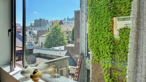Vista desde una ventana con cielo despejado, pared de ladrillo cubierta de hiedra verde y un edificio con tejado inclinado al fondo.