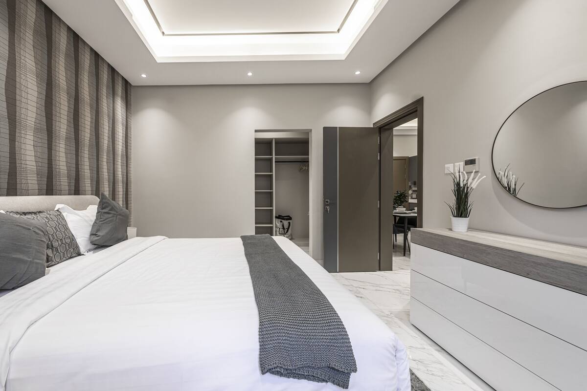 A modern bedroom with a skylight, a bed with grey bedding, a grey headboard, a white dresser, and a bathroom visible through an open door.