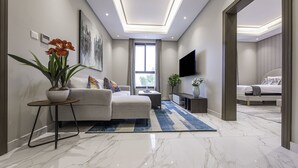 Ein modernes Wohnzimmer mit Sofa, Couchtisch und Fernseher. Ein großes Fenster und eine Tür zu einem anderen Raum sind vorhanden.