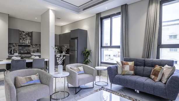 Ein modernes Wohnzimmer mit einem grauen Sofa, zwei Sesseln, einem Couchtisch und einer Küche im Hintergrund.