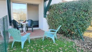 Une terrasse avec deux chaises bleu clair et une petite table, une haie verte et une porte vitrée donnant sur un espace intérieur.