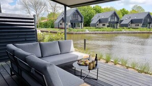A modern outdoor seating area with a grey sofa and a small wooden table, overlooking a calm water body and a row of contemporary houses. - Water Villa in Balk With Private Jetty (Balk)
