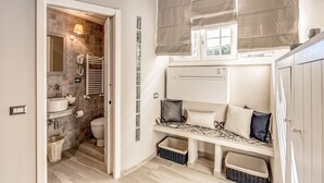 A bathroom with a sink, toilet, and shower. There is a bench with pillows and a basket.
