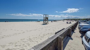 Una playa con un puesto de socorristas de madera, una fila de coches aparcados y un cielo azul despejado.