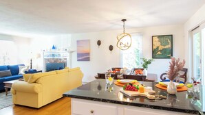 Una sala de estar moderna con un sofá, un comedor con una mesa y sillas, una isla de cocina con un bol de fruta, y un ventanal grande.