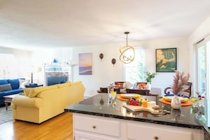 Un salon moderne avec un canapé, un coin repas comprenant une table et des chaises, un îlot de cuisine avec un panier de fruits et une grande fenêtre.
