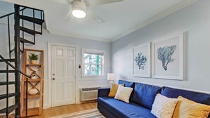 Uma sala de estar com um sofá azul, um tapete listrado, uma estante de madeira com uma planta, uma porta branca e uma janela com persianas.