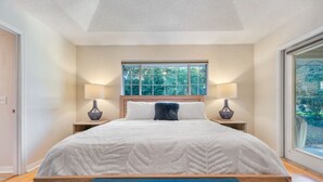 Um quarto com uma cama grande, duas luminárias de cabeceira, uma janela com vista para árvores e um banco azul estofado.