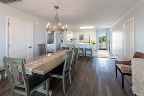 Un espace repas avec une longue table, des chaises, un lustre et une cuisine en arrière-plan.