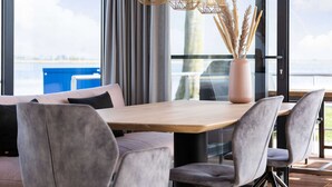 Un'area pranzo moderna con tavolo in legno e sedie rivestite in grigio, un vaso con piante essiccate e vista sul mare attraverso ampie finestre.