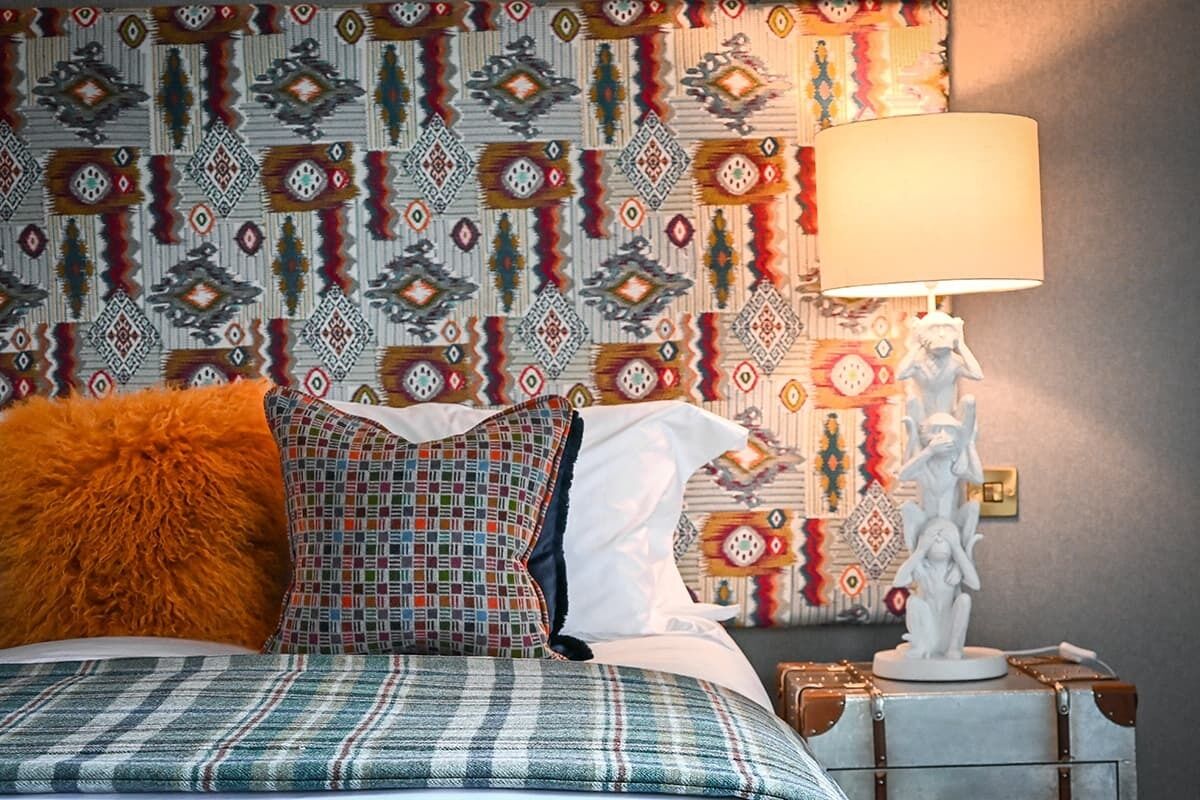 Una cama con ropa de cama escocesa, una almohada estampada y una almohada naranja suave. En la mesita de noche hay una lámpara y una estatua decorativa. El papel tapiz de la pared muestra un diseño geométrico.