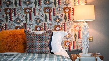 A bed with plaid bedding, a patterned pillow, and a fluffy orange pillow. There is a lamp on a nightstand with a decorative statue. The wall behind has a geometric patterned wallpaper.