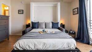 Ein ordentlich bezogenes Bett mit Kissen, ein Nachttisch mit Lampe, ein gerahmtes Bild an der Wand und ein Fenster mit Vorhängen.