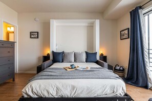 A neatly made bed with pillows, a bedside table with a lamp, a framed picture on the wall, and a window with curtains.