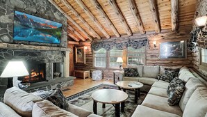 A cozy log cabin interior with a stone fireplace, a television mounted on the wall, and a comfortable seating area with a sofa and two round tables. - Spencer View (Waterfront on Moosehead Lake with Mountain Views) (Greenville)