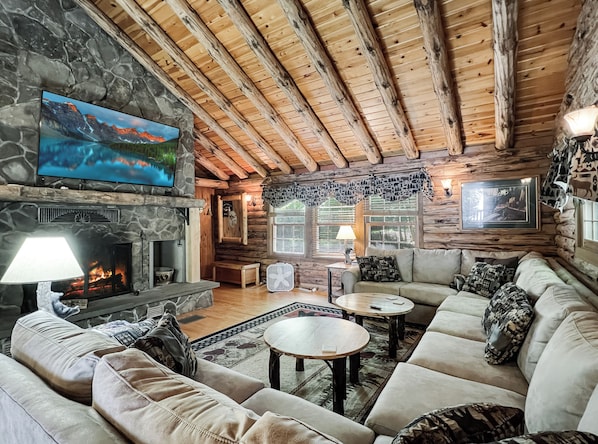 A cozy log cabin interior with a stone fireplace, a television mounted on the wall, and a comfortable seating area with a sofa and two round tables.