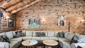 A log cabin interior with a sectional sofa, two round wooden tables, patterned cushions, a window with curtains, and wall-mounted lights. - Spencer View (Waterfront on Moosehead Lake with Mountain Views) (Greenville)