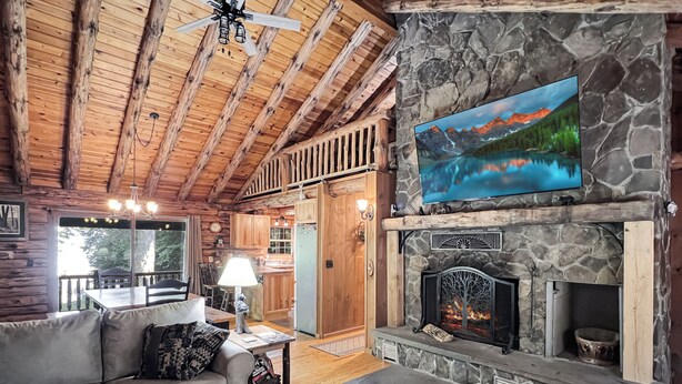 A cozy cabin interior with a stone fireplace, wooden ceiling beams, a dining area, and a large window overlooking the outdoors. - Spencer View (Waterfront on Moosehead Lake with Mountain Views) (Greenville)