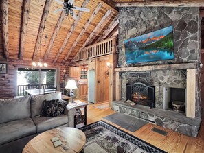 L’intérieur chaleureux d’un chalet avec une cheminée en pierre, des poutres en bois au plafond, un coin repas et une grande fenêtre donnant sur l’extérieur.