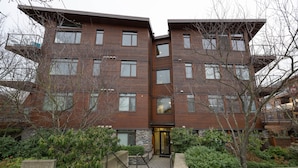 A multi-story building with a modern architectural design, featuring a central entrance with a glass door and a small outdoor area with steps and greenery. - Mt. Tolmie View-Spacious, Bright NE Facing Two Bedroom Condo with Den by Victoria Prime! (Victoria)
