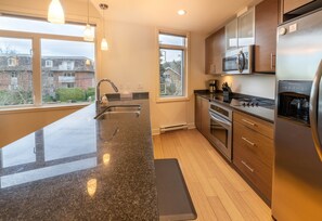 A modern kitchen with granite countertops, stainless steel appliances, and large windows offering views of a residential area. - Mt. Tolmie View-Spacious, Bright NE Facing Two Bedroom Condo with Den by Victoria Prime! (Victoria)